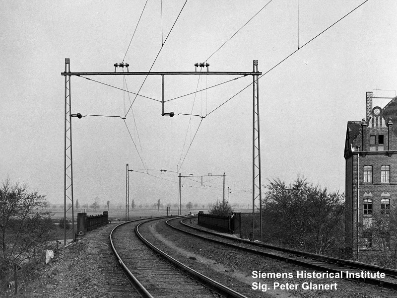 Einheitsfahrleitung im Bogen über eine Brücke. Rechts ein Haus. Im Hintergrund flaches Land mit Feldern.